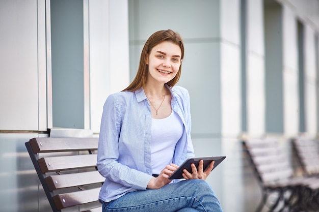 A young girl is sitting on a bench with a tablet in her hands and smiling.