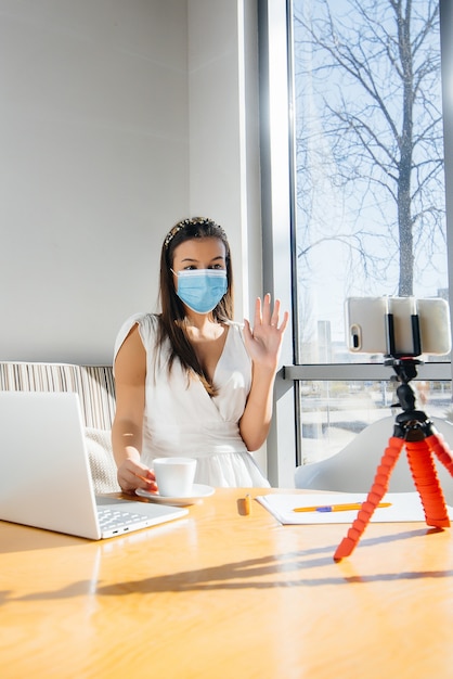 A young girl is sitting in a cafe in a mask and leads a video blog. Communication to the camera.