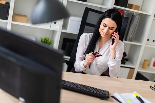 A young girl is sitting at a table, holding a bank card in her hand and talking on the phone.