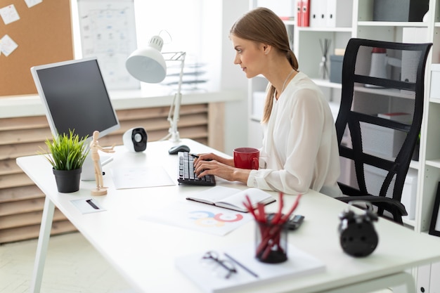 A young girl is sitting at a table in the office, holding a red cup in her hand and working at the computer.