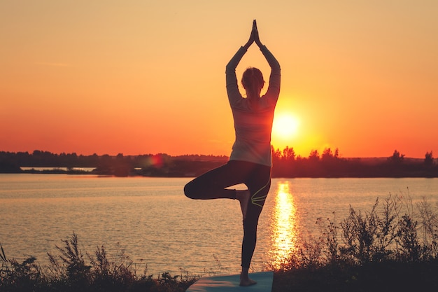 A young girl is standing on the lake at sunset, doing yoga