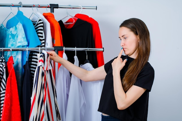 Young girl is thinking by holding colorful blouse on clothes background