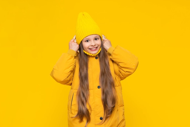 A young girl is warming herself in a warm autumn jacket A little girl in a down jacket and a hat wraps herself in clothes Children's stylish fashionable clothes Yellow isolated background