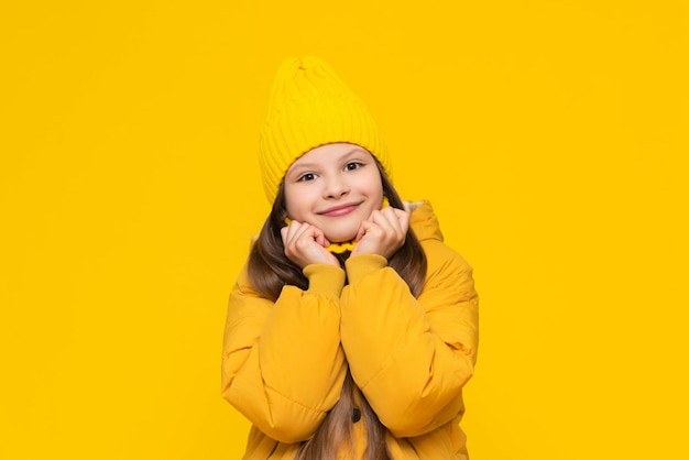 A young girl is warming herself in a warm autumn jacket A little girl in a down jacket and a hat wraps herself in clothes Children's stylish fashionable clothes Yellow isolated background