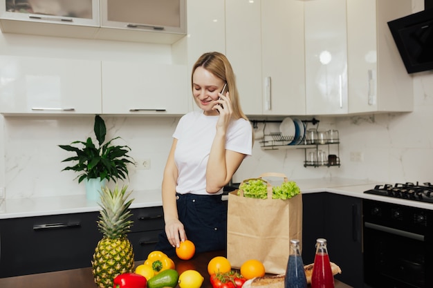 Young girl at kitchen holding phone and looking at vegetables and fruits on table.