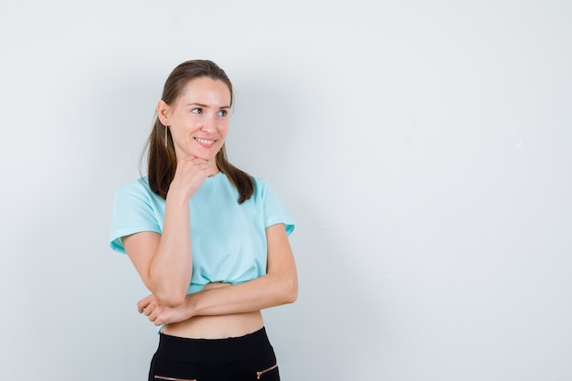 Young girl leaning chin on hand, standing sideways in turquoise t-shirt, pants and looking curious .