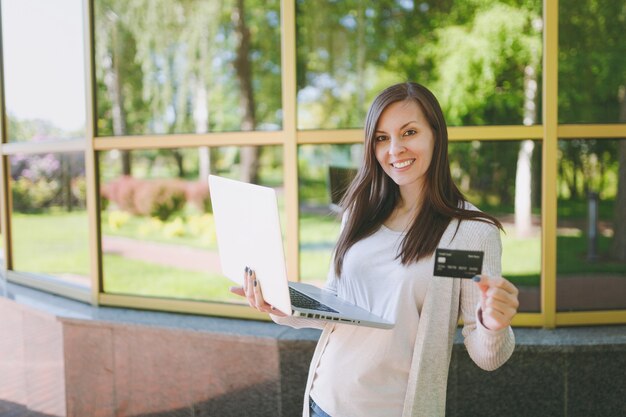 Young girl in light casual clothes holding credit card. Woman working on modern laptop computer near mirror building with tree reflection in street outdoors. Mobile Office. Freelance business concept.