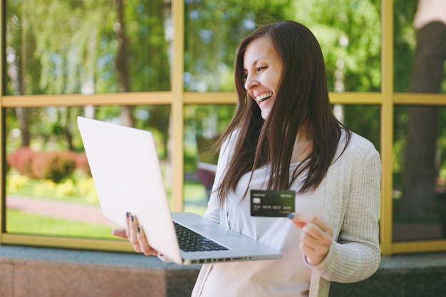 Young girl in light casual clothes holding credit card. Woman working on modern laptop computer near mirror building with tree reflection in street outdoors. Mobile Office. Freelance business concept.