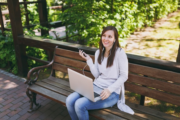 Young girl listen music in mobile phone with earphones. Woman sitting on bench working on modern laptop pc computer in city park in street outdoors on nature. Mobile Office. Freelance business concept