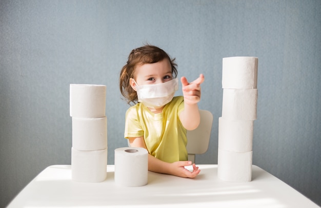 A young girl in a mask is sitting at a table, a girl in a white mask with toilet paper