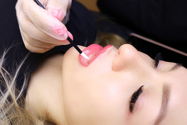 A young girl model is lying after the procedure of permanent lip makeup, the master rubs the oil with a brush on the lips of model