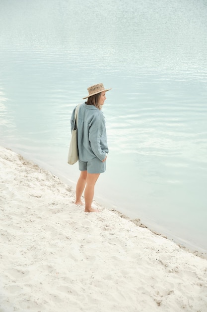 Young girl in natural linen clothes and a straw hat barefoot on a white sand beach holding a linen