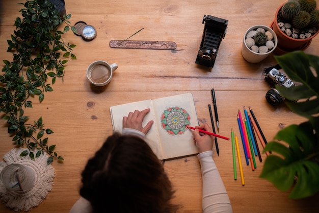 A young girl painting a mandala with pencils of many colors, on a wooden table. She has brushes, incense, cameras, and a large window. Top view.