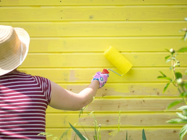 Young girl paints a wooden wall with a roller with yellow paint