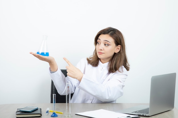 A young girl pharmacist in white coat pointing at glass bottle with liquid .