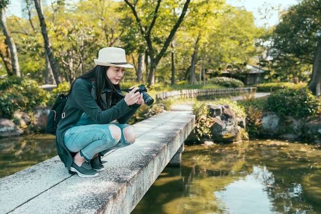 young girl photographer kneeling down on stone bridge and taking photo of pond in natural park in summer time. woman backpacker with camera on walkway across lake in forest while travel tokyo japan.