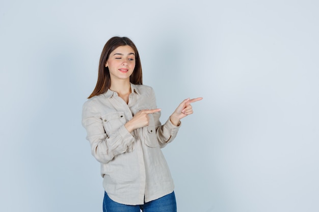 Young girl pointing right with index fingers in beige shirt, jeans and looking cute. front view.