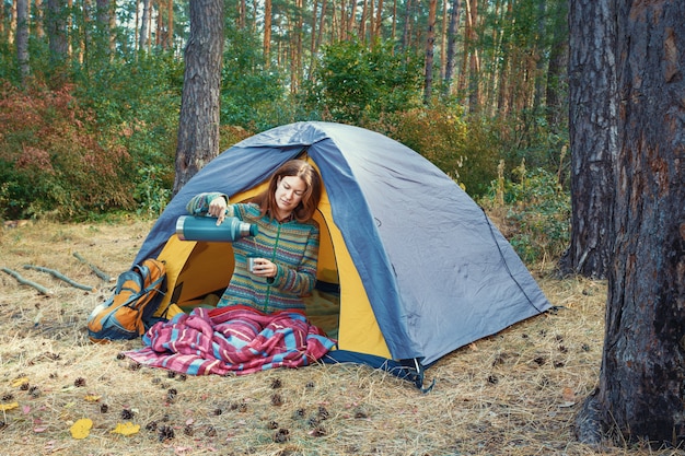 Young girl pouring tea in cup from thermos outdoors in camping tent