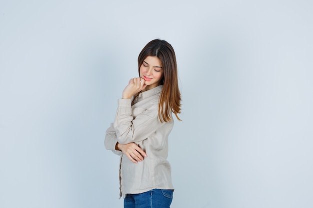 Young girl propping chin on hand, looking down in beige shirt, jeans and looking focused , front view.