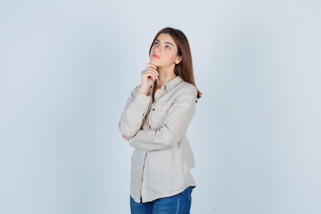 Young girl propping chin on hand, thinking about something in beige shirt, jeans and looking pensive. front view.