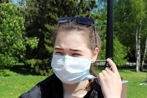 A young girl puts on a medical protective mask from viruses outside on a warm bright sunny day in a park during a pandemic.