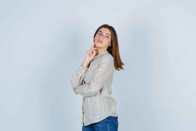 Young girl putting index finger on jowl, looking away in beige shirt, jeans and looking charming. front view.