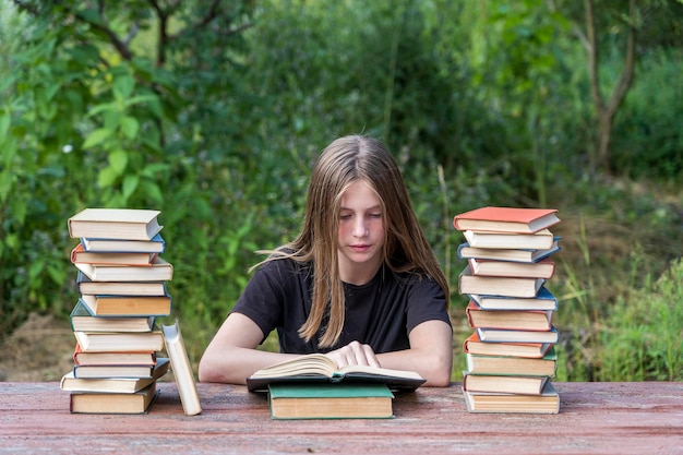 Young girl reading a book in the garden at a wooden table with a stack of books