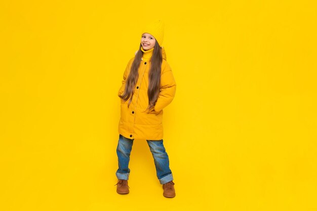 Photo a young girl rejoices at the onset of autumn warm clothes for children a child in an orange down jacket and hat on a yellow isolated background in the studio