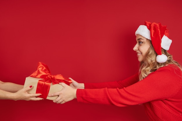 A young girl in a santa hat holds out her hands with a Christmas gift on a red background