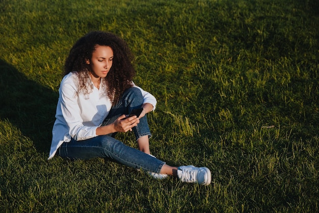 A young girl sits on the green grass in the park and uses social networks using a mobile phone