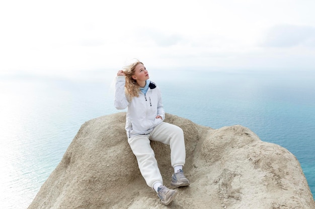 A young girl sits on a mountain top high above the sea on a sunny day Cute blonde in a white jacket and sweatpants Active lifestyle tourism and travel