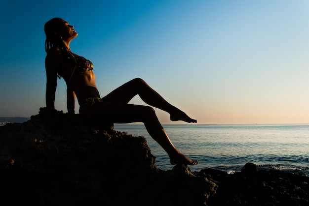 A young girl sits on a stone and enjoys the view of the sea and the sunrise