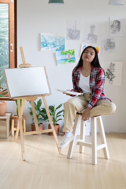 Young girl sitting on a chair with easel for drawing hold Color palette and brush in the room