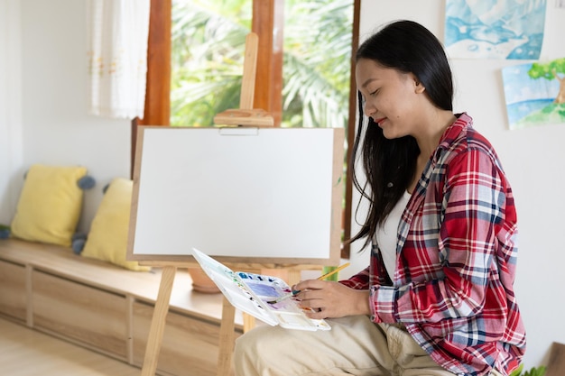 Young girl sitting on a chair with easel for drawing hold Color palette and brush in the room