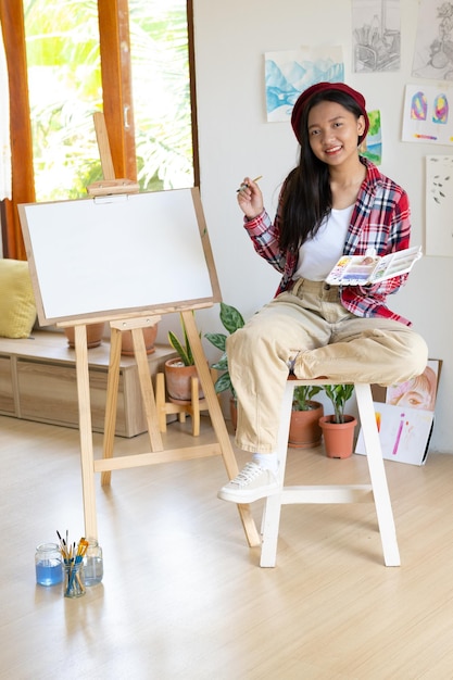 Young girl sitting on a chair with easel for drawing hold Color palette and brush in the room