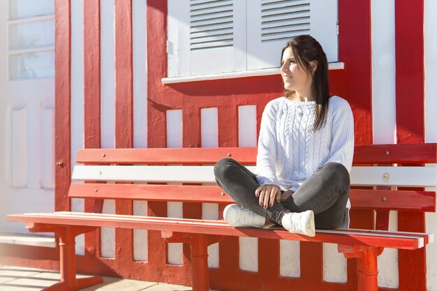 Young girl sitting crossed legged on a bench with a red and white striped background wall