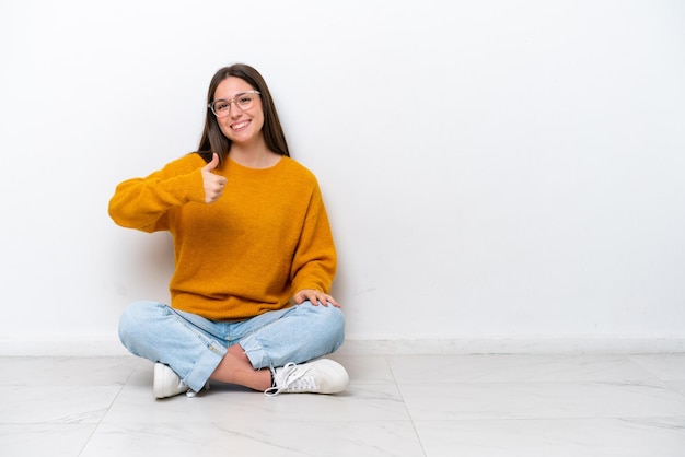 Young girl sitting on the floor isolated on white background giving a thumbs up gesture