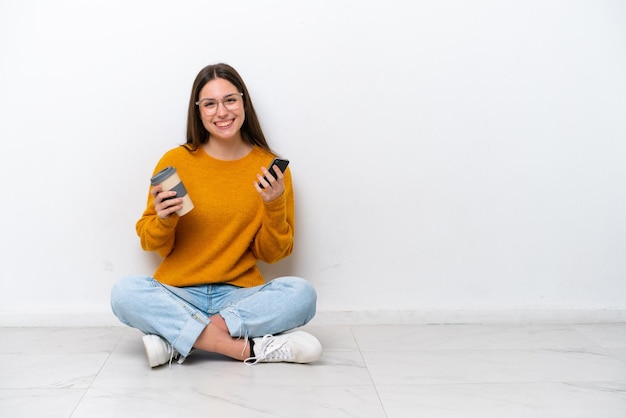 Young girl sitting on the floor isolated on white background holding coffee to take away and a mobile