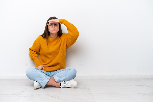 Young girl sitting on the floor isolated on white background looking far away with hand to look something