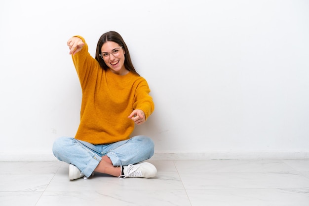 Young girl sitting on the floor isolated on white background points finger at you while smiling