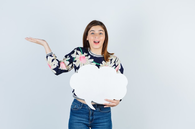 Young girl spreading palm in floral blouse, jeans and looking cheerful , front view.