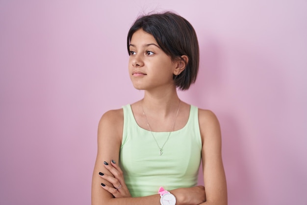 Young girl standing over pink background looking to the side with arms crossed convinced and confident