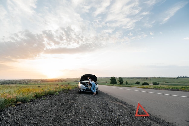 A young girl stands near a broken car in the middle of the highway during sunset and tries to repair it Troubleshooting the problem Waiting for help Car service Car breakdown on road