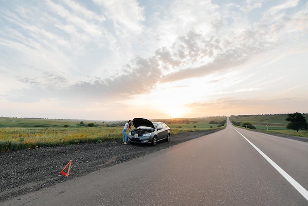 A young girl stands near a broken car in the middle of the highway during sunset and tries to repair it Troubleshooting the problem Waiting for help Car service Car breakdown on road