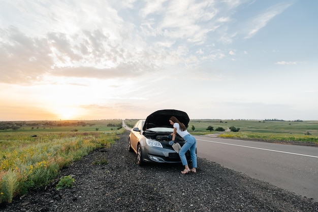 A young girl stands near a broken car in the middle of the highway during sunset and tries to repair it Troubleshooting the problem Waiting for help Car service Car breakdown on road