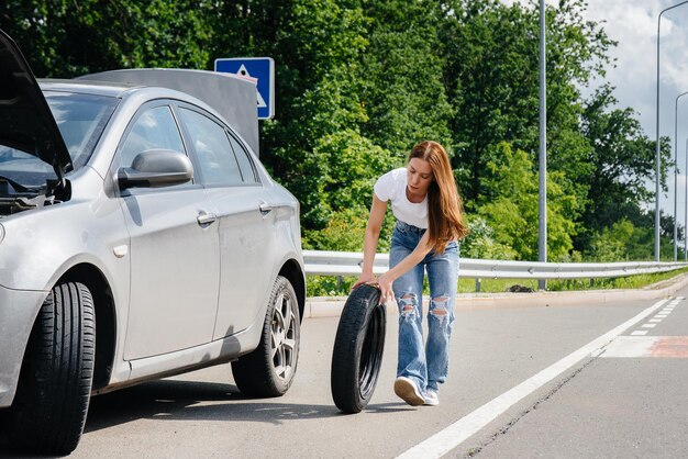 Photo a young girl stands near a broken car in the middle of the highway and tries to change a broken wheel on a hot sunny day. failure and breakdown of the car. waiting for help.