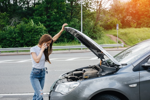 A young girl stands near a broken-down car in the middle of the highway and calls for help on the phone