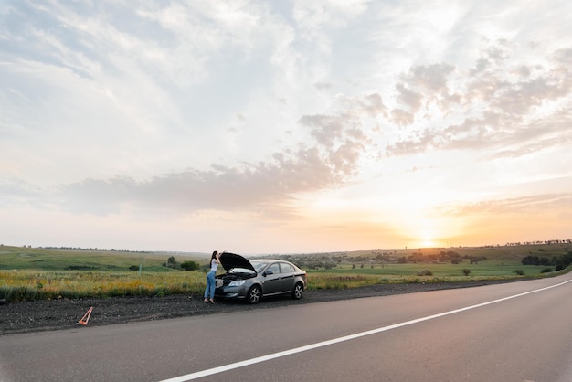 A young girl stands near a brokendown car in the middle of the highway during sunset and tries to call for help on the phone Waiting for help Car service Car breakdown on road