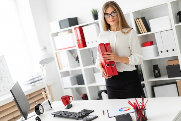 A young girl stands near a table in the office and holds a folder with documents.