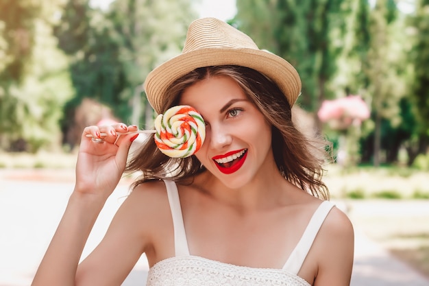 Young girl in a straw hat and a multi-colored lollipop walks in the park